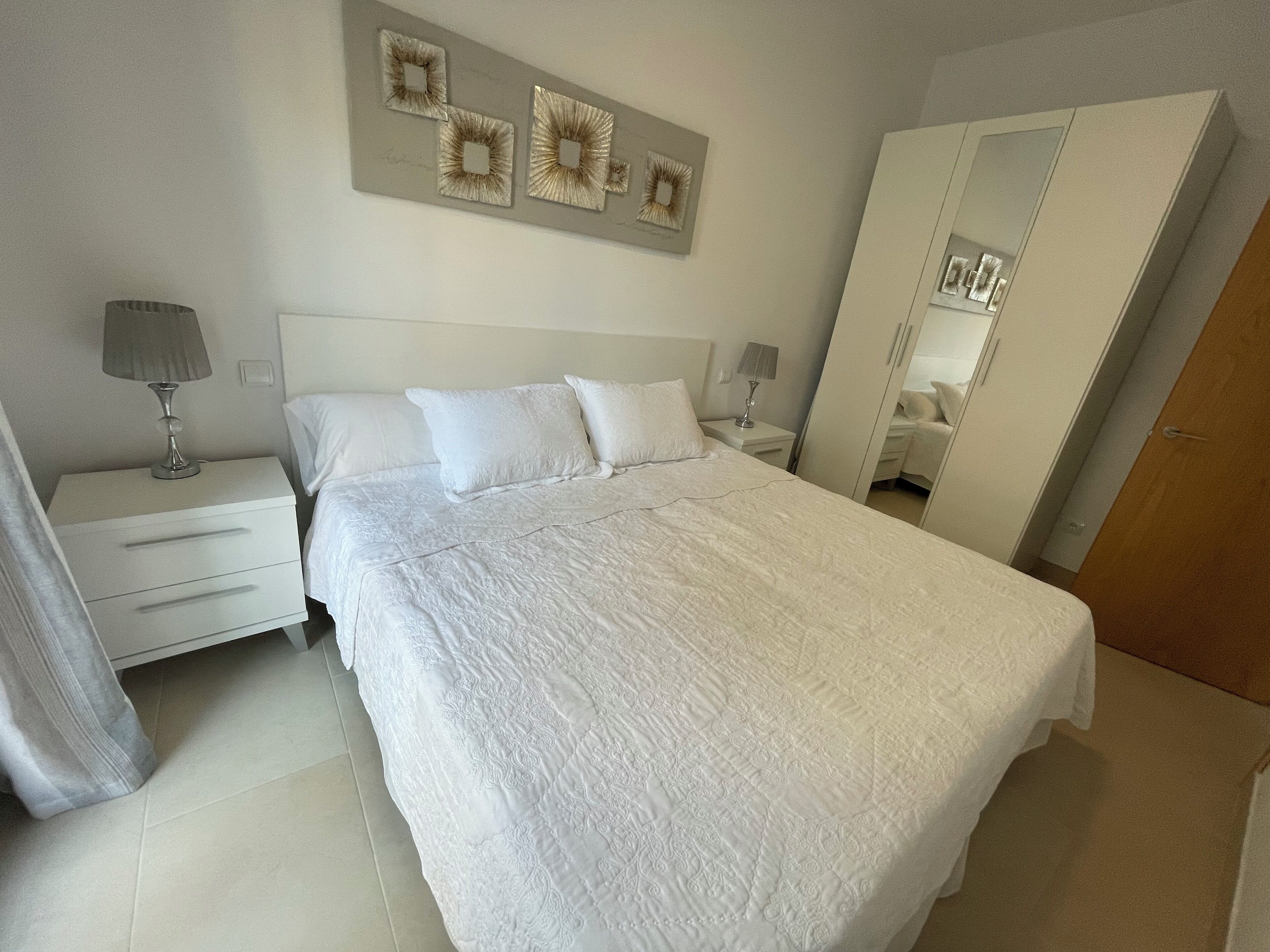 A neatly made bed with a white comforter and pillows, a bedside table with a lamp, a wall-mounted mirror, and a door to the right.