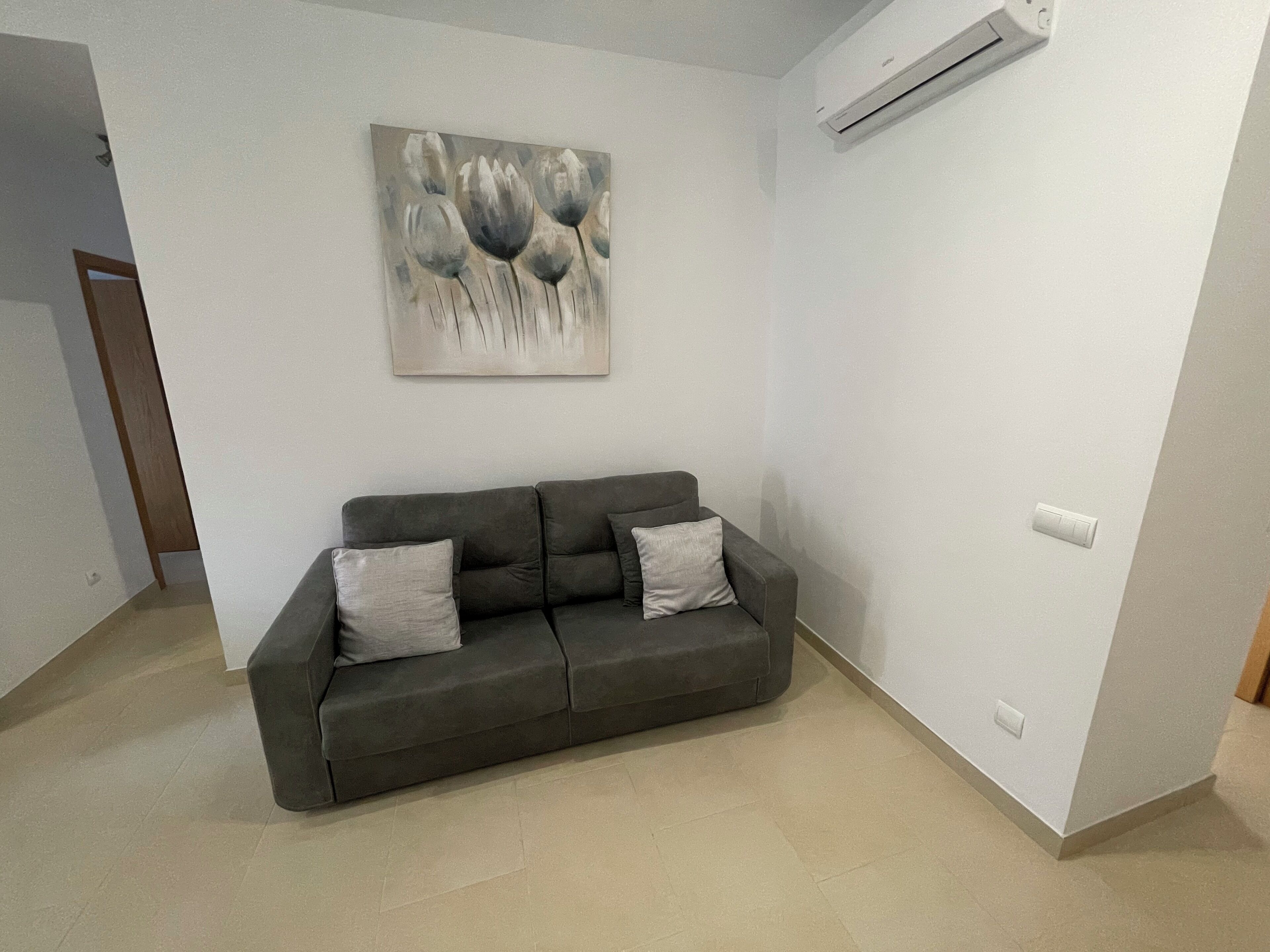 A modern living room with a grey sofa, two cushions, a wall-mounted air conditioner, and a minimalist abstract painting.