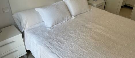 Una cama bien hecha con ropa blanca y almohadas, una mesita de noche con lámpara y una cómoda.