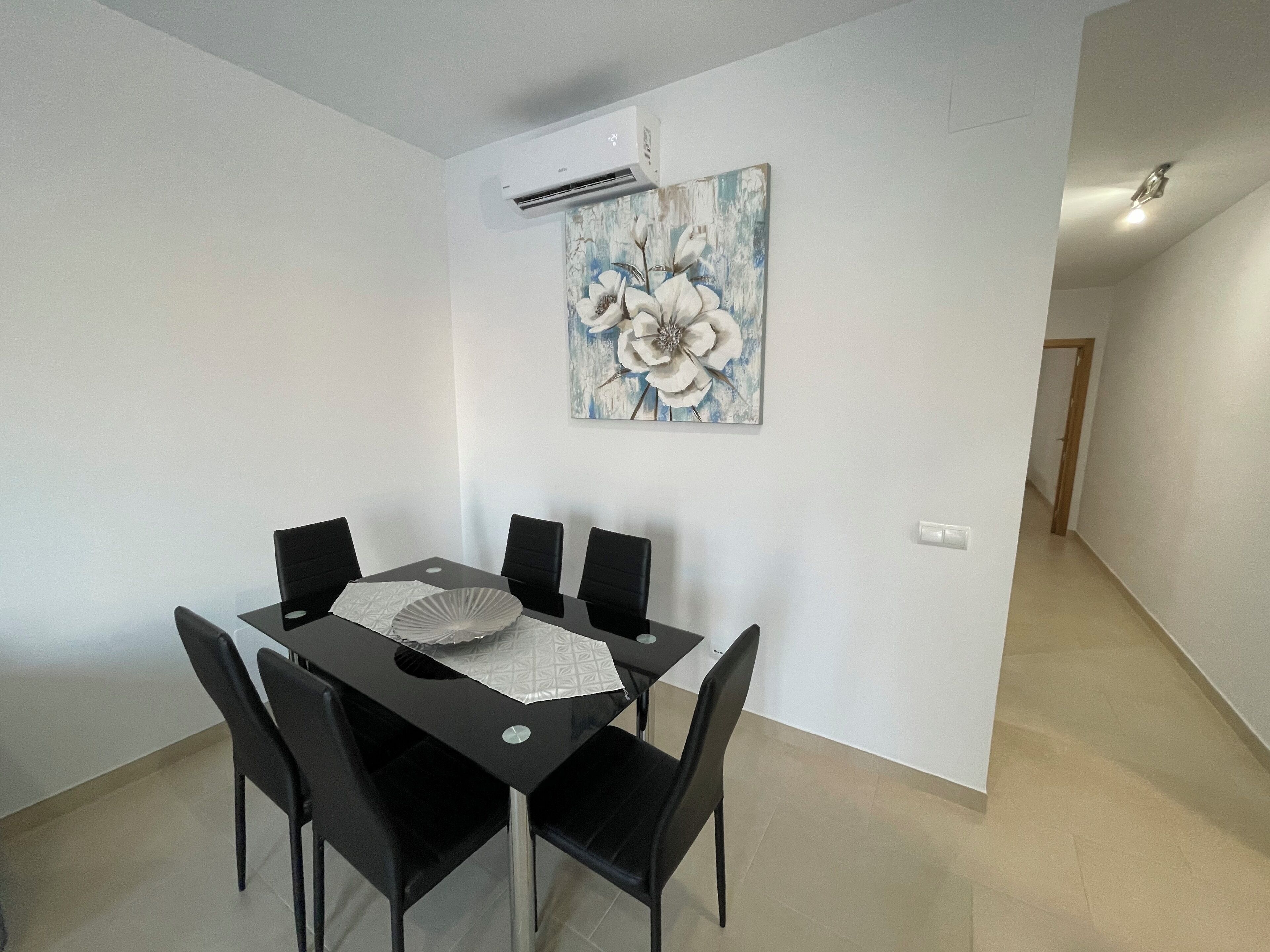 Una zona de menjador amb taula i cadires negres, una paret blanca amb un quadre floral i una unitat de aire condicionat.
