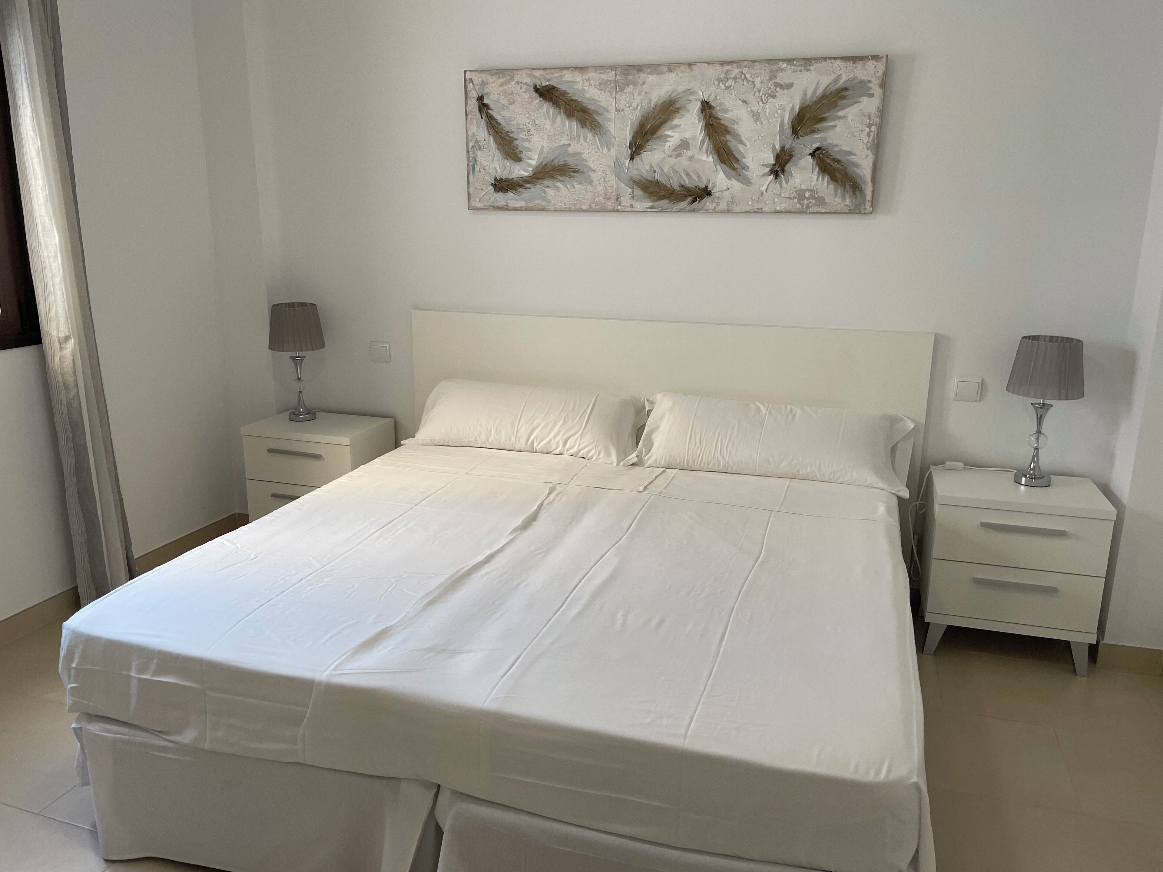A bedroom with a large bed, two bedside tables, a wall-mounted artwork, and a window with curtains.