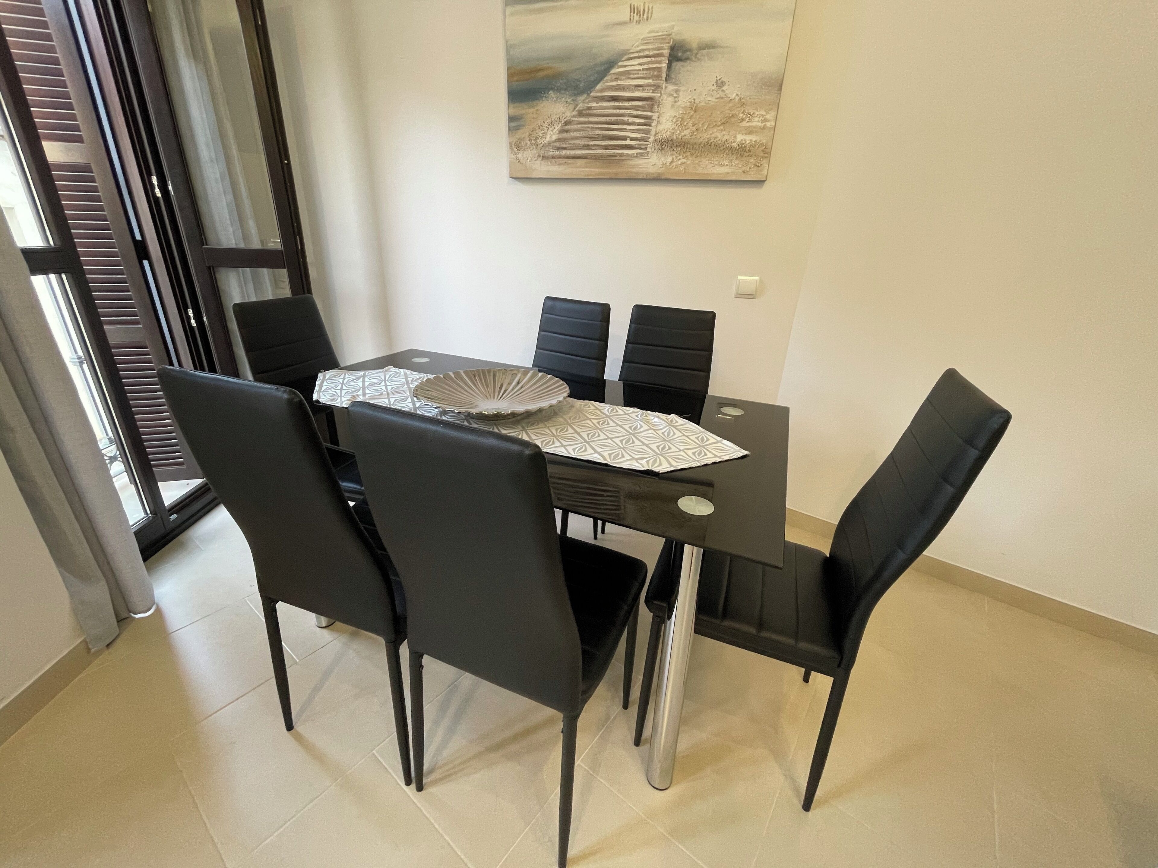 Un coin repas avec une table dressée pour quatre personnes, des chaises noires, une table en verre et un tableau accroché au mur.