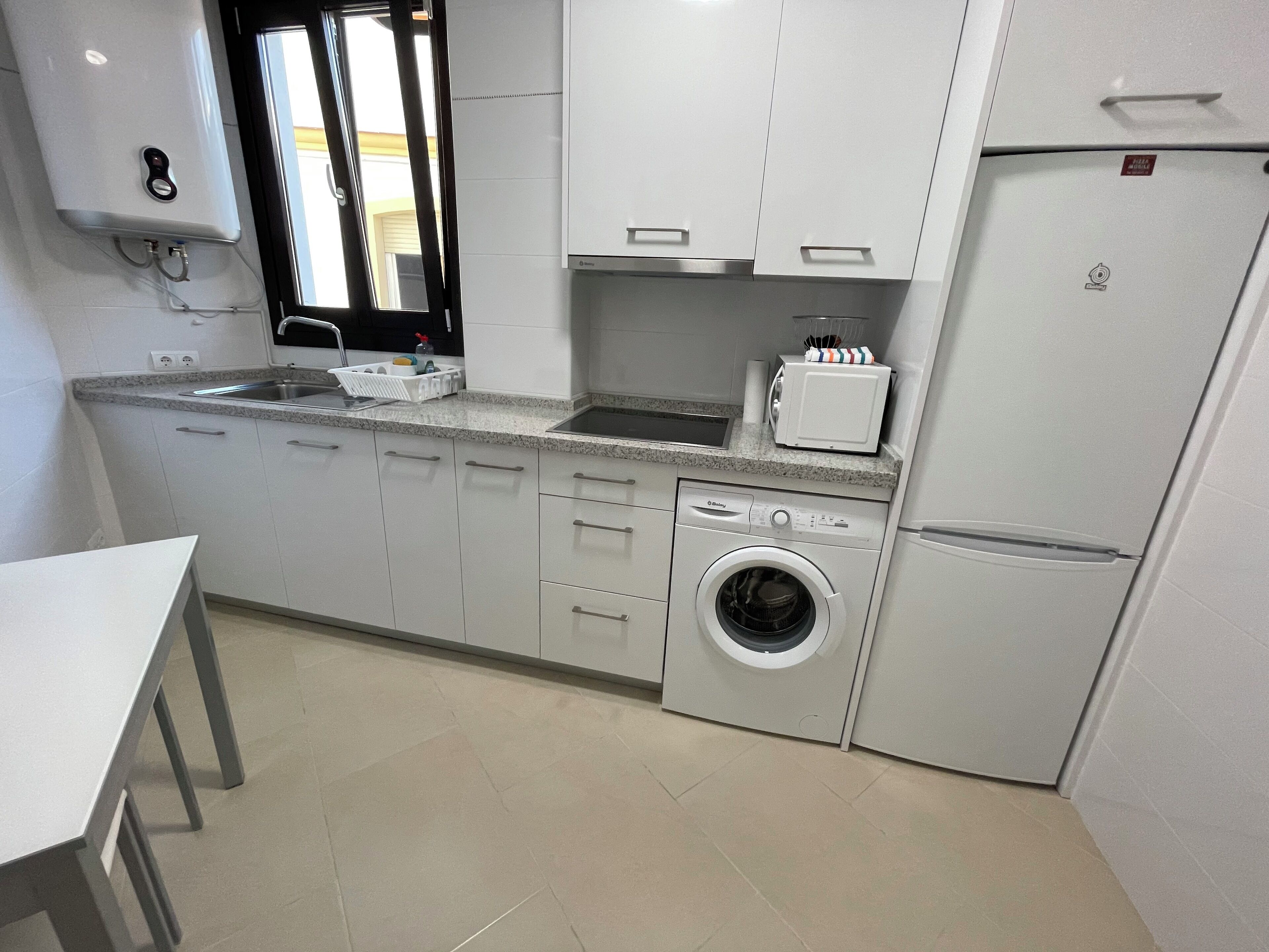 Uma cozinha compacta com armários brancos, máquina de lavar embutida, frigorífico e micro-ondas.