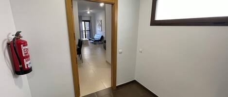 A hallway with a fire extinguisher mounted on the wall, a door leading to a room with a bed and a window, and a light fixture above the door.