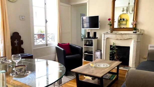 A living room with a glass table, a black armchair, a wooden coffee table, a fireplace, and a TV on the wall. - L'appartement Boulnois (Paris)