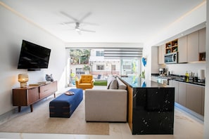 Un salon moderne avec un téléviseur à écran plat, un canapé, un fauteuil jaune, une table basse et un coin cuisine doté d’un plan de travail noir.