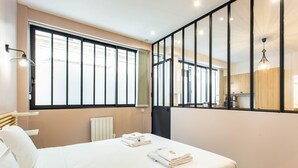 A hotel room with a large bed, a window with blinds, a radiator, and a view of the kitchen.
