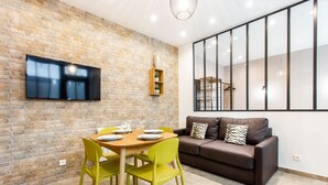 A modern living room with a brick wall, a flat-screen TV, a brown leather sofa, and a dining area with green chairs and a wooden table.