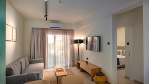 A modern living room with a sofa, a coffee table, a TV mounted on the wall, and a door leading to a bedroom. - Amore Protaras Apartments (Paralimni)