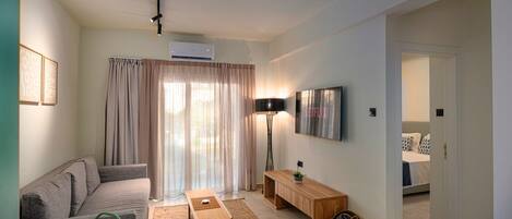 Una sala de estar moderna con un sofá, una mesa de centro, un televisor montado en la pared y una puerta que da a un dormitorio.