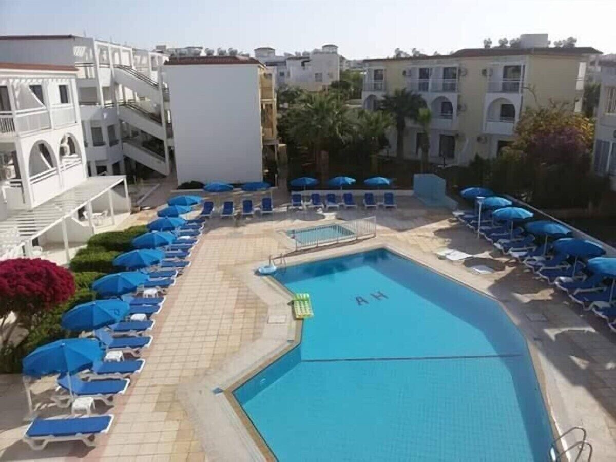 A swimming pool with blue umbrellas and lounge chairs, surrounded by buildings and palm trees.