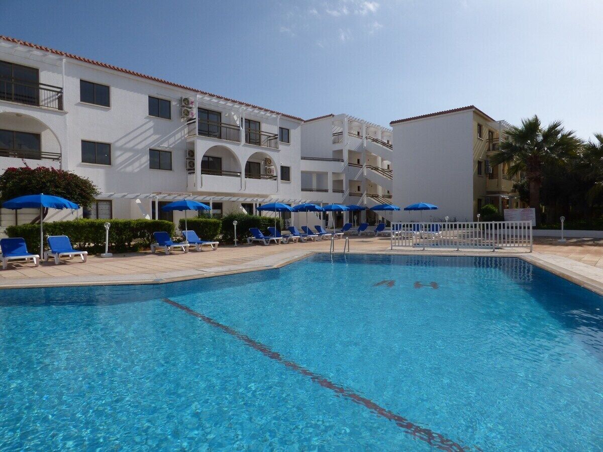 A swimming pool with blue water, surrounded by white buildings with balconies, and blue umbrellas.