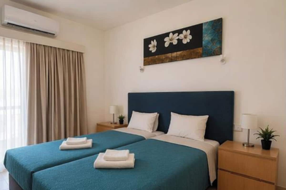A hotel room with two beds, a nightstand with a lamp, a wall-mounted air conditioner, and a painting above the beds.