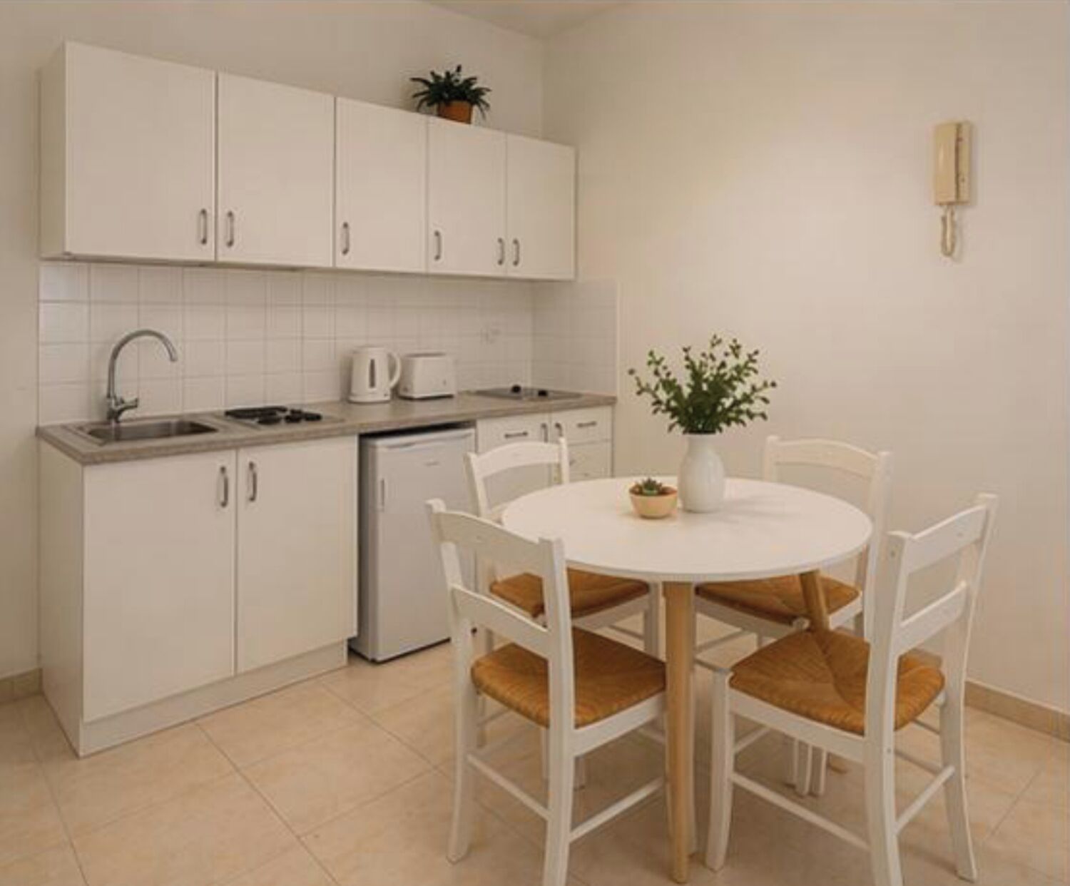 A kitchen with white cabinets, a sink, a toaster, and a small potted plant on the counter. There is a white dining table with four chairs and a vase with a plant on it.