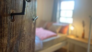 En trædør med metalgreb og nøglehul, der fører ind til et soveværelse med seng, puder og et natbord med lampe.
