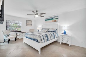 A bedroom with a white bed, a ceiling fan, a desk with a chair, a nightstand with a lamp, and a beach-themed painting on the wall. - NPR (Naples)