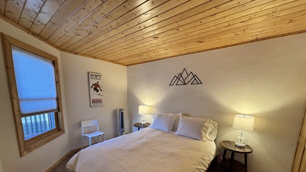 A bedroom with a wooden ceiling, a bed with white bedding, two bedside lamps, a chair, and a poster on the wall.