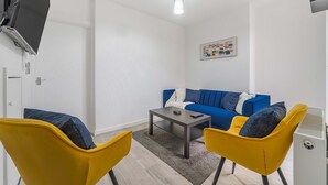 A modern living room with a blue sofa, yellow chairs, a coffee table, and a flat-screen TV mounted on the wall.