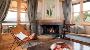 A cozy room with a fireplace, a wooden bench, a cushioned chair, and a table with a fruit bowl.