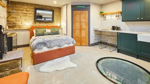 A modern bedroom with a stone wall, a wooden door labeled "RAVENSCAR," a bed with a fur throw, a small kitchen area with a countertop and cabinets, and a unique glass floor feature.