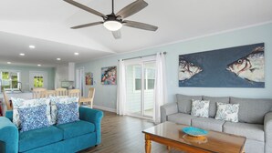 A spacious living area with a blue sofa, a grey sofa, and a wooden coffee table. There is a ceiling fan, a painting of fish on the wall, and a kitchen in the background. - Spectacular views from this 2nd Row East side Duplex - New for 2025 Shared Pool (Emerald Isle)