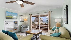 Ein Wohnzimmer mit Deckenventilator, einem Holztisch, beigen Sofas und Blick auf einen Balkon mit Geländer.