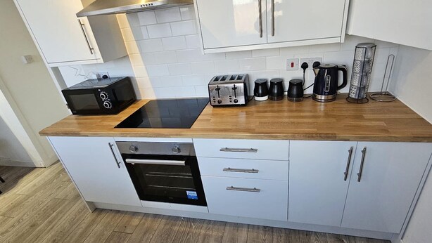 A compact kitchen with white cabinets, a black induction cooktop, a built-in oven, and a coffee maker. - Cozy 3-bed House in Dagenham (Dagenham)