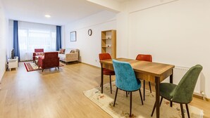Ein modernes Wohnzimmer mit einem Holzesstisch, roten und blauen Stühlen, einem Sofa und einem Bücherregal.