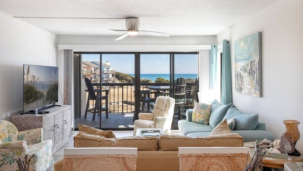 A spacious living room with a large TV, comfortable seating, and a balcony with a view of the ocean. - Sound of the Sea West 115 - Almost Oceanfront 2 BR 2 BA Condo (Emerald Isle)