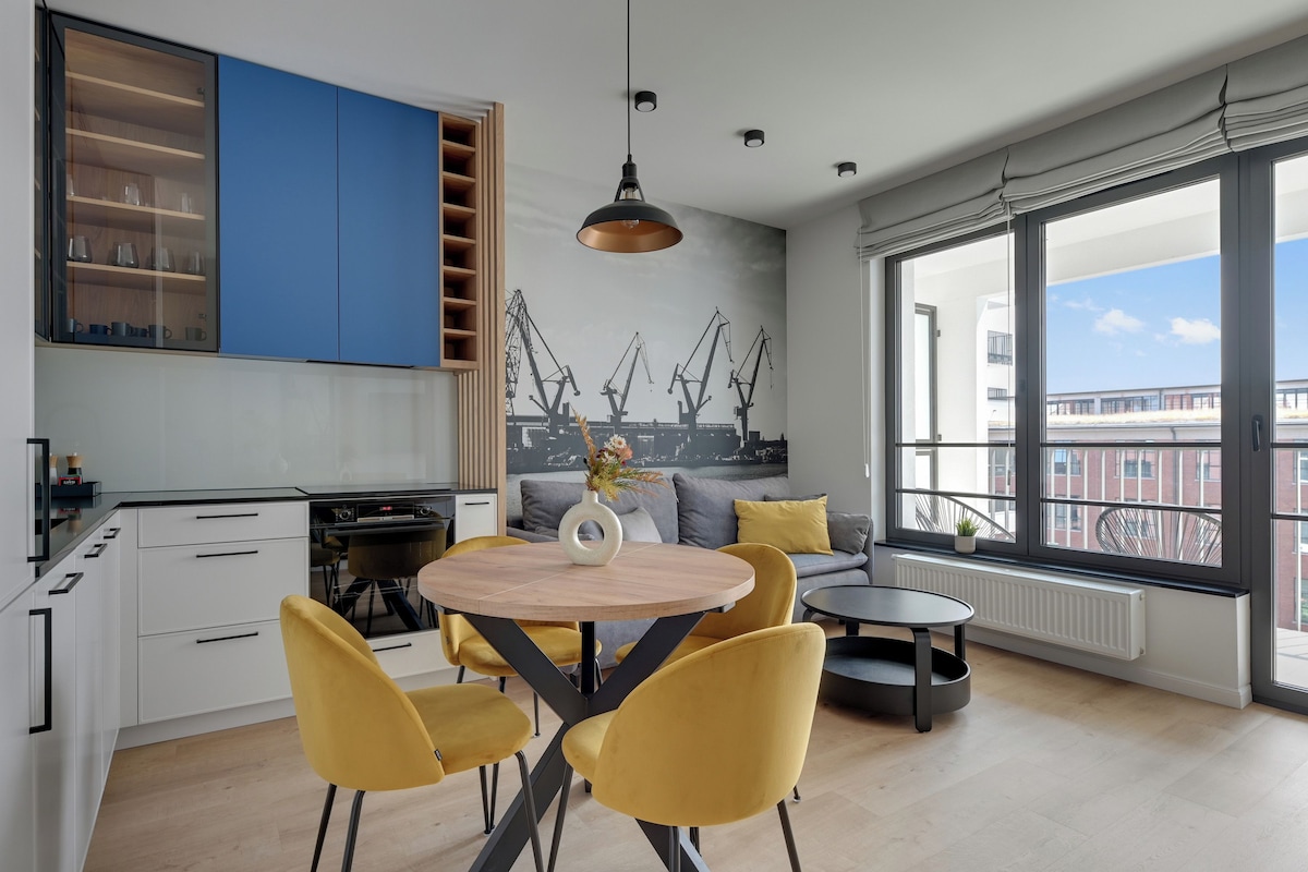 A modern kitchen with white cabinets, a blue accent wall, and a dining area with yellow chairs and a round table.