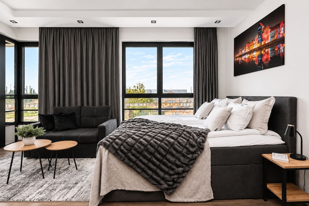 A modern bedroom with a bed, a sofa, a coffee table, and large windows offering a city view.