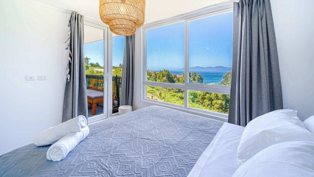 Un dormitorio con un ventanal grande, una cama, una lámpara colgante y vistas a la vegetación y al mar.