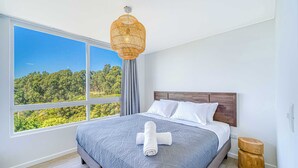 Un dormitorio con un ventanal grande, una cama con colcha gris, almohadas blancas, cabecera de madera y una lámpara de fibra.