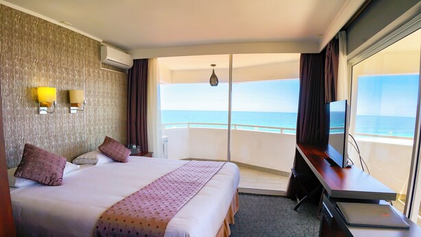 A hotel room with a large bed, a desk with a laptop, a TV, and a view of the ocean.