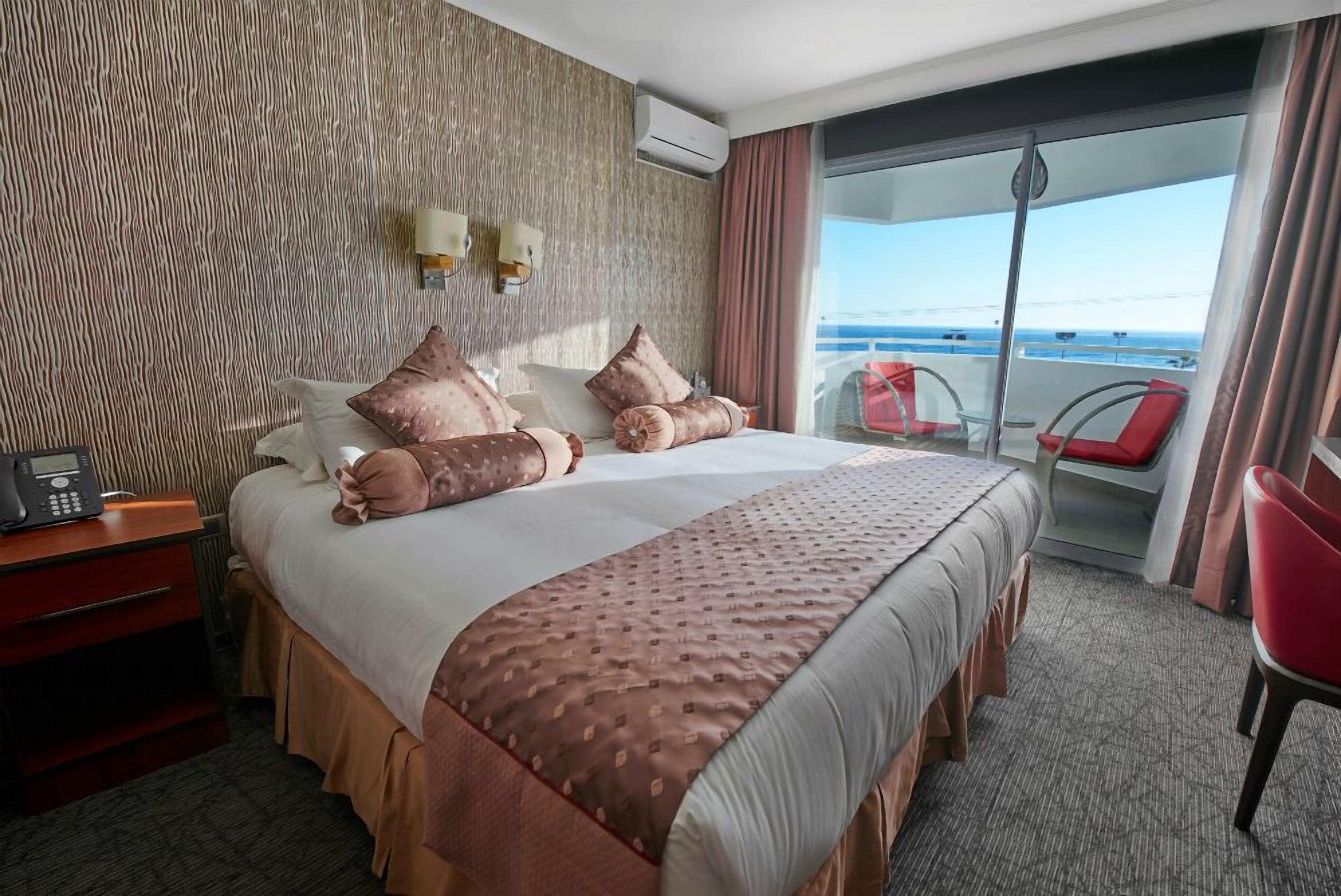 A hotel room with a large bed, a bedside table with a phone, a view of the balcony with red chairs, and a textured wall.