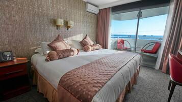 Habitación de hotel con una cama grande, una mesita de noche con teléfono, vista al balcón con sillas rojas y una pared con textura.