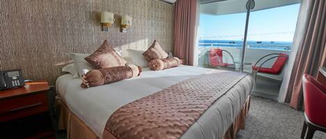 A hotel room with a large bed, a bedside table with a phone, a view of the balcony with red chairs, and a textured wall.