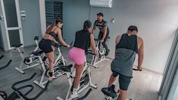 En gruppe mennesker træner på spinningcykler i et fitnesscenter med grå vægge og en dør til venstre.