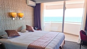 A hotel room with a large bed, a nightstand, a desk, and a chair. There is a window with a view of the ocean.