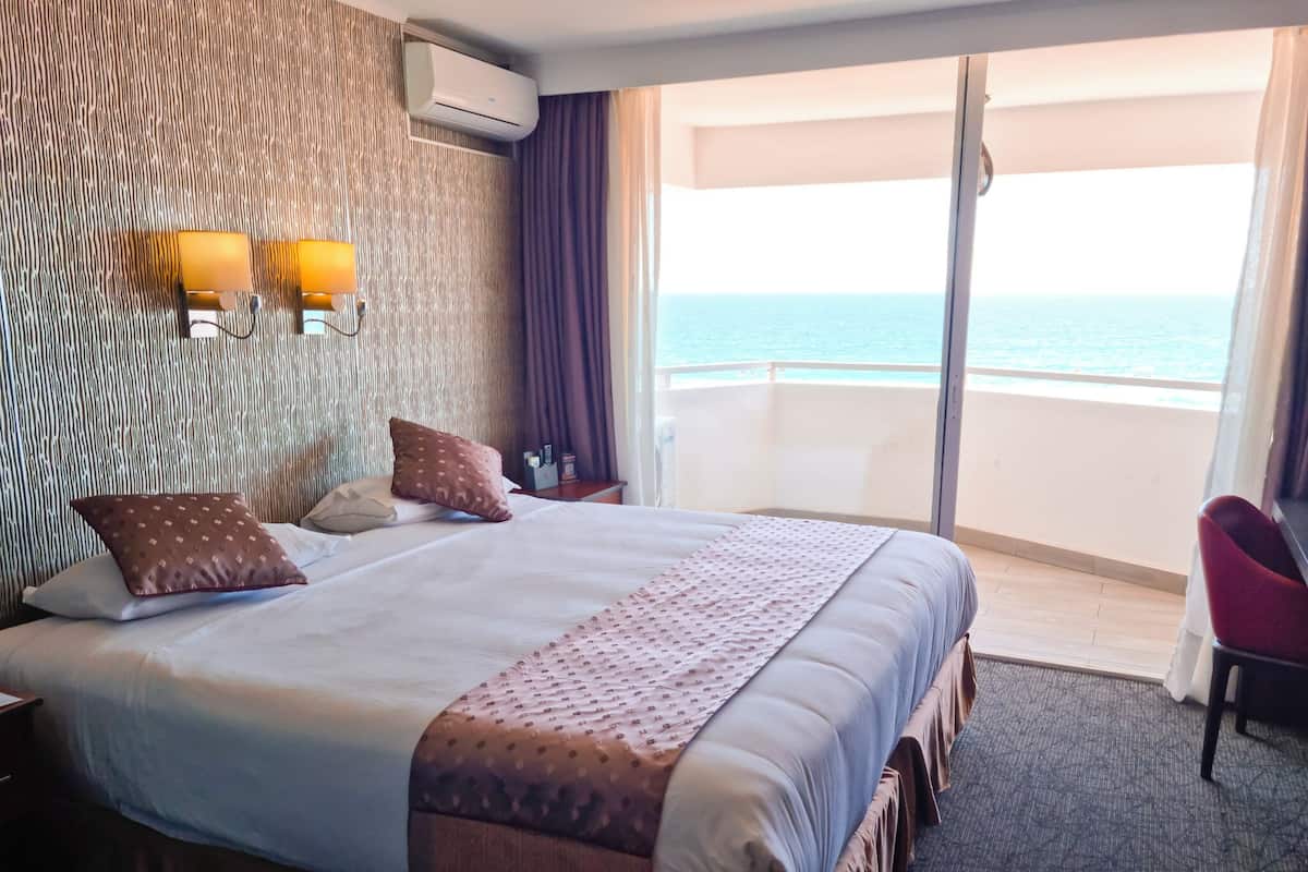 A hotel room with a large bed, a nightstand, a desk, and a chair. There is a window with a view of the ocean.