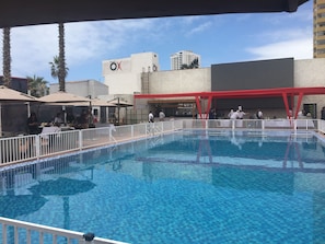 Una piscina con borde de baldosas, rodeada por una cerca blanca, y un edificio con un cartel que dice "ESPACIO" al fondo.
