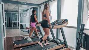 Two people walking on treadmills in a gym with a large window and a mirror.