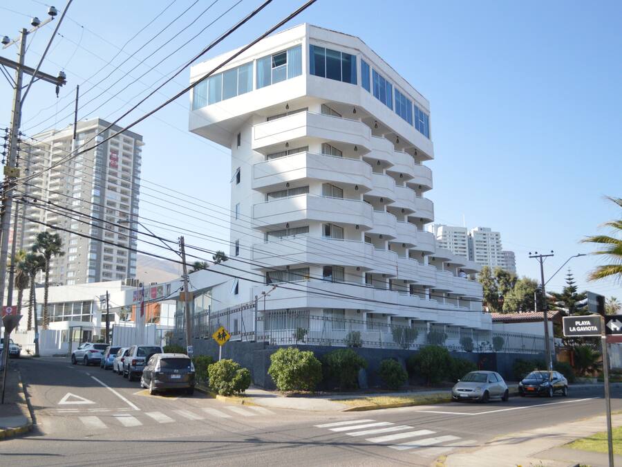 Un edificio moderno de varios pisos con balcones, un paso de peatones y coches estacionados.