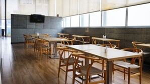 A spacious indoor dining area with wooden tables and chairs, large windows, and a flat-screen TV mounted on the wall.