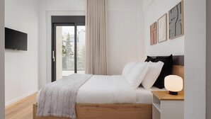 A modern bedroom with a wooden bed, a bedside table with a lamp, a TV mounted on the wall, and a window with curtains. - Glyfada Experience Residence (Glyfada)