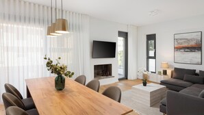 A modern living room with a wooden dining table, comfortable seating, a flat-screen TV, a fireplace, and a large window with sheer curtains. - Glyfada Experience Residence (Glyfada)