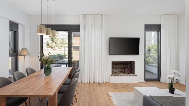 A modern living room with a wooden dining table, grey chairs, a flat-screen TV mounted on the wall, a fireplace, and large windows with white curtains. - Glyfada Experience Residence (Glyfada)