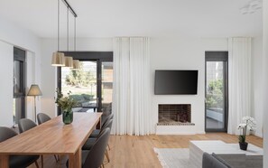 A modern living room with a wooden dining table, grey chairs, a flat-screen TV mounted on the wall, a fireplace, and large windows with white curtains. - Glyfada Experience Residence (Glyfada)