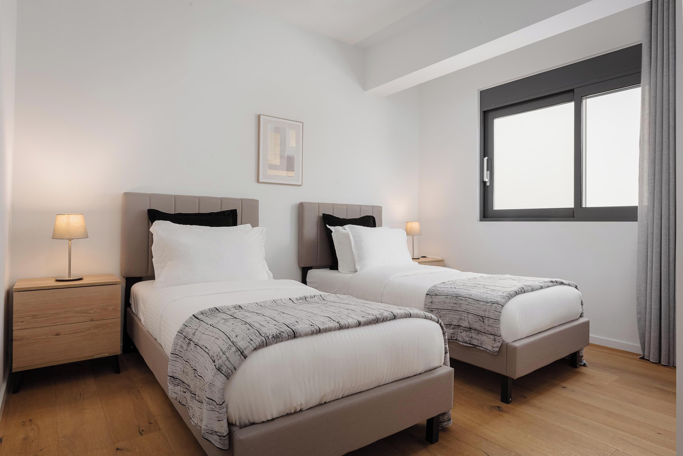 A hotel room with two beds, a nightstand with a lamp, a framed picture on the wall, and a window with curtains.