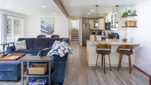 A modern living room with a blue sofa, a coffee table, and a kitchen area with a bar and stools.